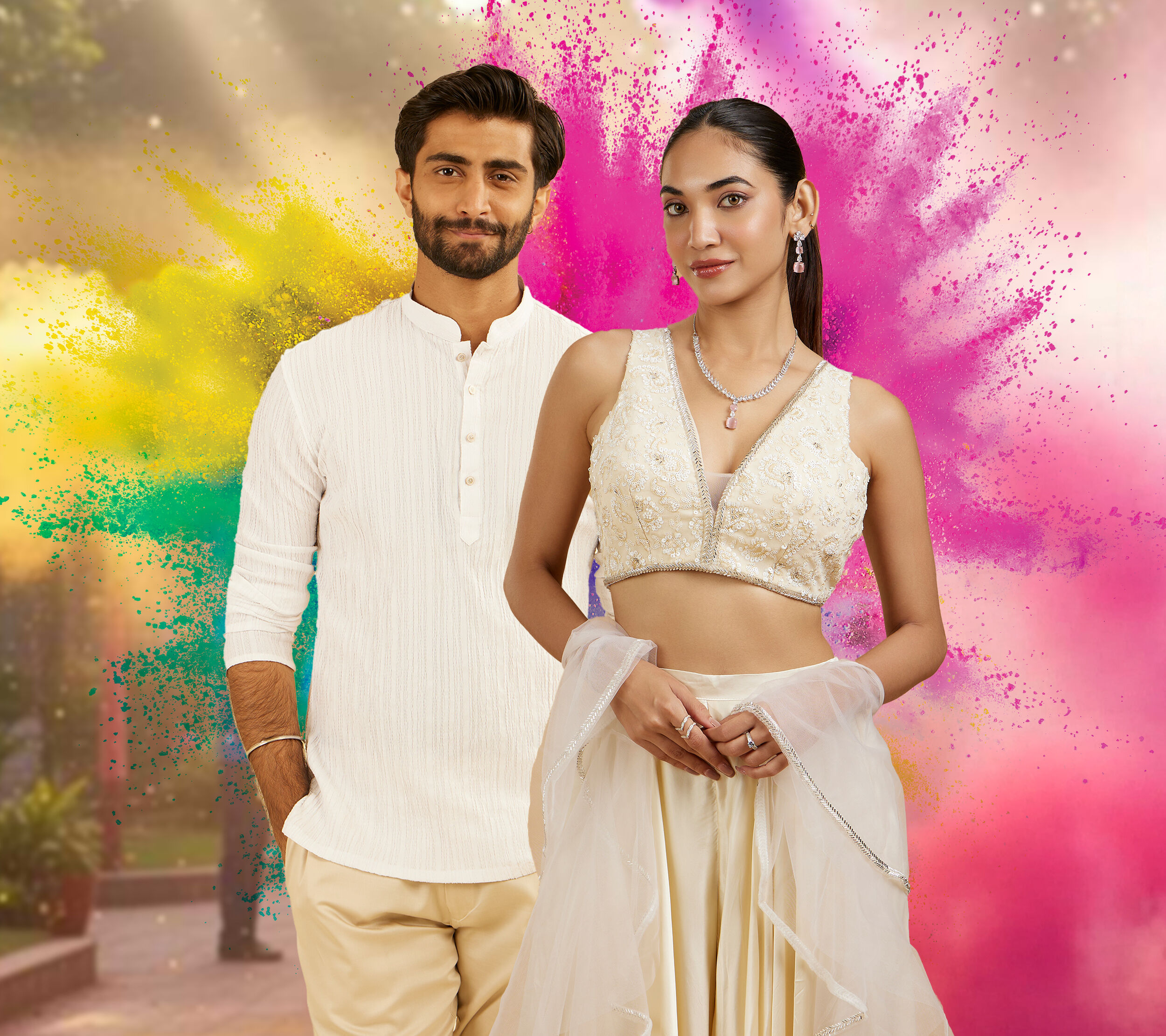 A stylish couple posing in festive traditional outfits during Holi, standing in front of vibrant splashes of pink, yellow, and green colors in the background.